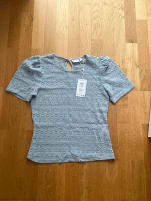 Ljusblå spetsblus - Söt ljusblå blus med kort puffärm och rund halsringning. Blusen har ett fint spetsmönster över hela plagget och en liten knappdetalj i nacken. Perfekt för dig som gillar romantiska detaljer och en feminin look.