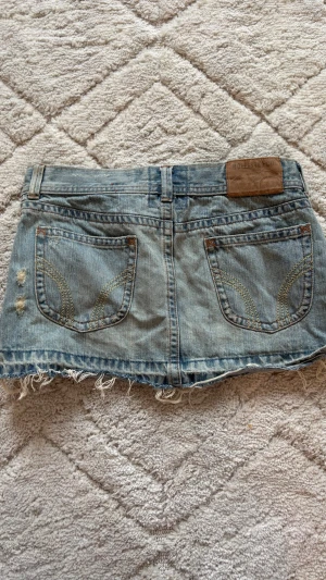 Ljusblå vintage jeanskjol från Hollister - Trendig ljusblå jeanskjol från Hollister med slitna detaljer och fransig nederkant. Kjolen har två bakfickor med dekorativa sömmar och klassisk jeanslook. Perfekt för en avslappnad och cool stil.