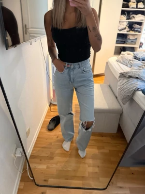 Ljusblå raka jeans med hål på knäna - Säljer ett par ljusblå jeans med raka ben och slitningar på båda knäna. Jeansen har hög midja och klassisk femficksdesign. Perfekta för en avslappnad och trendig look.