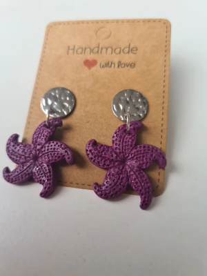 Lilaglittriga sjöstjärnor - Unika handgjorda örhängen med lila, glittriga sjöstjärneformade hängen och silverfärgade, hamrade knappar. Örhängena är gjorda för hand av mig i polymerlera. 