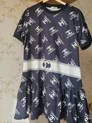 Svart kort klänning med logomönster - Snygg svart kort klänning med vit logotypmönster över hela plagget. Klänningen har rund halsringning, korta ärmar och volang nedtill. Tillverkad i ett mjukt och lätt material, perfekt för dig som gillar statement-plagg.