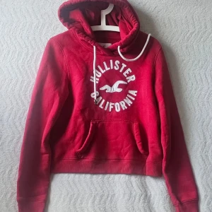 Röd hoodie från Hollister - Snygg röd hoodie från Hollister med vit broderad text och logga på bröstet. Klassisk känguruficka framtill, ribbade muddar och justerbar huva med vita snören. Mjuk insida för extra komfort. Perfekt för en avslappnad och trendig look.