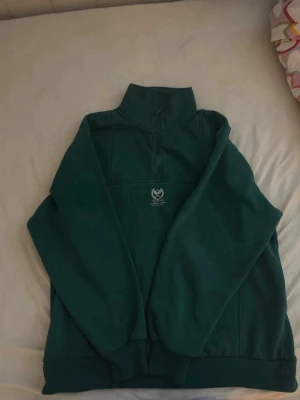Mörkgrön halfzip hoodie - Mörkgrön hoodie med halv dragkedja och hög krage. Tröjan har ett litet vitt tryck på bröstet med text och symbol. Tillverkad i mjukt sweatshirtmaterial och har ribbade muddar vid ärmslut och nederkant. Perfekt för en chill och avslappnad stil. Köpte den i Turkiet från defacto