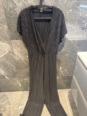 Plisserad svart jumpsuit med v-ringning - Snygg svart plisserad byxdress från Lindex i storlek M.   Endast använd en gång och i mycket bra skick. Omlottdesign framtill med liten knapp vid urringningen och band i midjan som kan knytas för en fin passform. Vida ben och elegant modell, perfekt till fest, middag eller andra tillfällen.  Kan skicka flera bilder om det önskas eller frågor om mått osv 😊