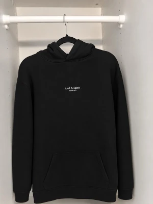 Svart hoodie Axel Arigato - Snygg svart hoodie från Axel Arigato med vit logga broderad på bröstet. Klassisk modell med huva och stor magficka framtill. Tillverkad i mjukt material som känns skönt mot huden. Perfekt för en avslappnad och trendig look.