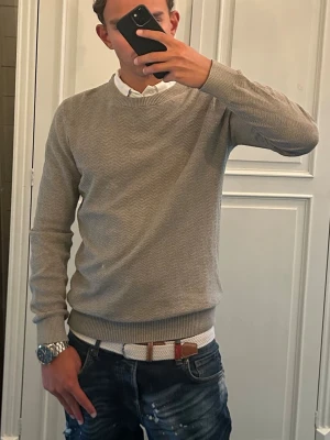 Stickad tröja Dressmann - Säljer en stilren grå stickad tröja från Dressmann med diskret sicksackmönster. Tröjan har rund halsringning, ribbade muddar vid ärmslut och nederkant. Perfekt att bära över en skjorta för en clean look.