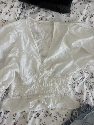 Vit blus med v-ringning och puffärm - Säljer en vit blus med djup v-ringning och bred smock i midjan. Blusen har puffiga långärmar och lätt volang nedtill. Perfekt för dig som gillar en romantisk och luftig stil. Materialet känns tunt och svalt, troligen bomull eller bomullsblandning.