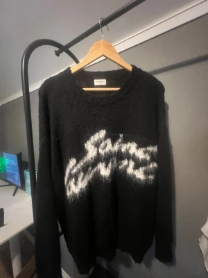 Svart Saint Laurent stickad tröja - Säljer en svart stickad tröja från Saint Laurent med vit, fluffig logga framtill. Tröjan har rund halsringning och långa ärmar. Materialet ser ut att vara mohairmix som ger en mjuk och lite lurvig känsla. Perfekt för dig som gillar streetstyle och exklusiva märken.