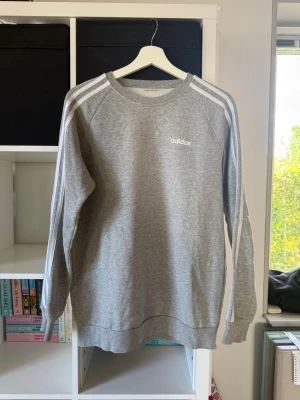 Grå sweatshirt från Adidas - Klassisk grå sweatshirt från Adidas med vita ränder längs ärmarna och Adidas-logga på bröstet. Tröjan har rund halsringning, långa ärmar och ribbade muddar. Tyvärr finns det ingen lapp ja men jag skulle tro storlek S/M 