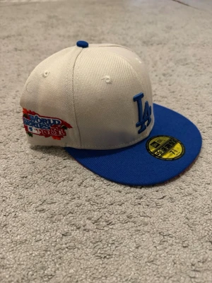 LA Dodgers keps med blå skärm - Snygg keps med vit krona och blå skärm, broderad LA-logga framtill och patch på sidan med texten 'World Series 1981'. Klassisk 59FIFTY-modell med rak skärm och klistermärke kvar på ovansidan. Perfekt för dig som gillar streetwear och sportig stil.