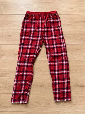 Rödrutiga pyjamasbyxor med snörning - Mysiga pyjamasbyxor i rött rutigt mönster med svarta och vita detaljer. Byxorna har resår i midjan och snörning för extra komfort. Tillverkade i mjukt tyg, perfekta för chillkvällar hemma. Liten gul logga på vänster sida.