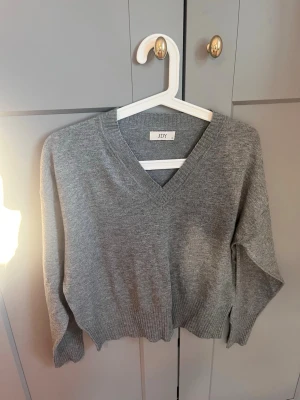 Grå stickad tröja med v-ringning JDY - En enkel och stilren grå stickad tröja från JDY med v-ringad hals och långa ärmar. Tröjan har ribbade muddar vid ärmslut och nederkant, vilket ger en snygg passform. Perfekt att matcha med jeans eller kjol för en chill look.