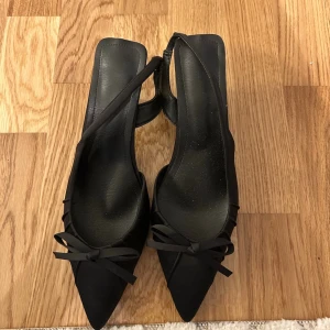 Svarta spetsiga pumps med rosett - Säljer ett par svarta pumps med spetsig tå och låg klack. Skorna har en elegant rosett framtill och öppen häl med rem. Materialet ser ut att vara matt tyg, vilket ger en stilren look. Perfekta för dig som gillar klassiska och trendiga skor.