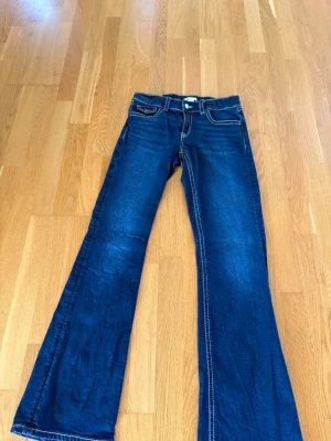 Mörkblå bootcut jeans med kontrastsömmar - Snygga mörkblå bootcut jeans med vita kontrastsömmar och klassiska fem fickor. Jeansen har utsvängda ben och detaljerade bakfickor med knapp. Aldrig använda då jag råka beställa i fel storlek.