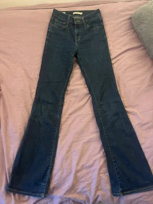 Mörkblå bootcut jeans - Säljer ett par mörkblå bootcut jeans med hög midja och klassisk femficksdesign. Jeansen har utsvängda ben och kontrastsömmar. Perfekta till sneakers eller boots för en snygg look.