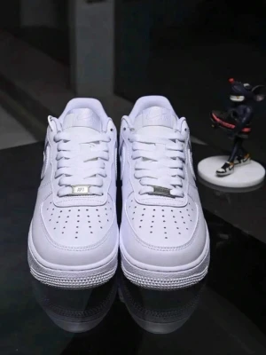 Vita klassiska sneakers - Stilrena vita sneakers från Nike med lågt skaft och perforerad tåbox för extra ventilation. Skorna har en ikonisk design med tydlig swoosh på sidan, vita skosnören och metallbricka med 'AF1' på snörningen. Tillverkade i slätt läder för en clean look.