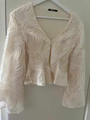 Krämvit meshkofta med struktur - Säljer en unik krämvit kofta från Gina Tricot i meshmaterial med swirl-struktur och transparenta partier. Koftan har vida ärmar och knäppning framtill. Perfekt för dig som vill sticka ut med en cool och trendig look.