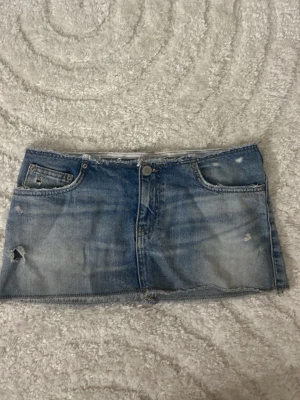 Blå jeanskjol från Pull&Bear - Säljer en blå jeanskjol från Pull&Bear med rå kant och slitna detaljer. Kjolen har klassiska fem fickor och knappgylf. Perfekt för dig som gillar en avslappnad och trendig stil.