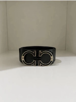 Svart skärp med guldspänne - Snyggt svart skärp med ett stort, iögonfallande guldspänne i unik design. Perfekt accessoar för att lyfta vilken outfit som helst. Skärpet är gjort i skinn och har en stilren look som passar till många olika stilar.