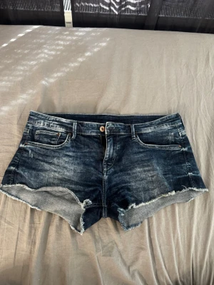 Mörkblå jeansshorts med rå kant - Säljer ett par mörkblå jeansshorts med slitna detaljer och rå, fransig kant. Shortsen har klassisk femficksdesign, knappgylf och en avslappnad passform. Perfekta för varma dagar och en chill stil. Storlek L ( vet ej vilket märke)