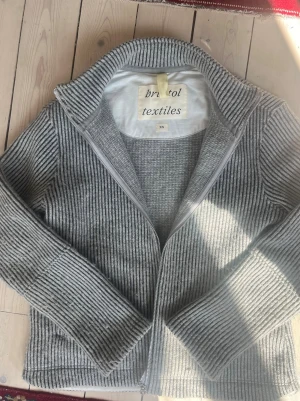 Grå ribbad ziptröja från bristol textiles - Nypris 1999kr.  Snygg grå ribbstickad ziptröja från bristol textiles i storlek XS. Tröjan har dragkedja framtill, hög krage och är tillverkad av delvis återvunna material. Perfekt för lager på lager och ger en clean look.