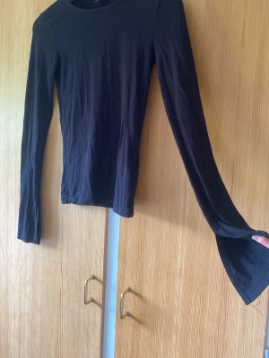 Svart långärmad topp med slits - En svart långärmad topp med slits vid ärmsluten. Toppen har en enkel, tight passform och är tillverkad i ett mjukt material som känns bekvämt mot huden. Perfekt att styla med jeans eller kjol för en clean look.