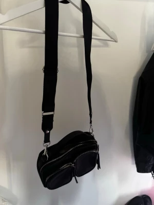 Svart crossbody handväska med dragkedjor - Snygg svart handväska i crossbody-modell med bred justerbar axelrem. Väskan har flera fack framtill med silverfärgade dragkedjor och är tillverkad i ett slätt konstlädermaterial. Invändigt finns ett ljust foder och praktiska innerfickor för småsaker.