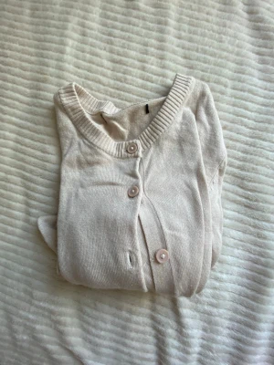 Ljusbeige tunn kofta med knappar - En stilren ljusbeige kofta med rund halsringning och långa ärmar. Koftan har ribbade muddar och knäpps med matchande knappar framtill. Perfekt att slänga över en topp för en enkel och clean look.