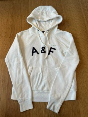 Vit hoodie från Abercrombie & Fitch - Snygg vit hoodie med stor huva och dragsko. På bröstet finns ett tydligt A&F-tryck i svart. Hoodien har en klassisk känguruficka framtill och är gjord i mjukt material, perfekt för en avslappnad stil.