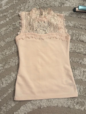 Beige spetsdetaljerad ärmlös topp - Supersöt beige topp med spets upptill och hög hals. Toppen är ärmlös och har fina spetsdetaljer både fram och bak, vilket ger en romantisk vibe. 