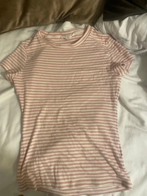 Rosa och vit randig t-shirt - Säljer en söt t-shirt med rosa och vita ränder. T-shirten har korta ärmar, rund halsringning och är gjord i ett mjukt bomullsmaterial. Perfekt för dig som gillar en enkel men fräsch stil.