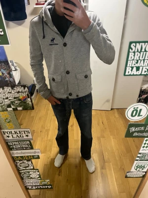 Grå hoodie från Abercrombie & Fitch - Snygg grå hoodie med knappar och huva från Abercrombie & Fitch. Liten logga på bröstet och text längs ena ärmen. Två fickor framtill med lock och ribbade muddar. Perfekt för en avslappnad och trendig stil. Storleken är mellan xs och s mått på sista bilden 