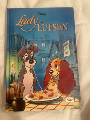 Lady och Lufsen Disney bok - En färgglad barnbok med hårda pärmar från Disney, 'Lady och Lufsen'. Omslaget visar de två hundarna Lady och Lufsen vid ett bord med spaghetti. Boken har illustrationer i blå, röd och gul ton och är skriven på svenska.