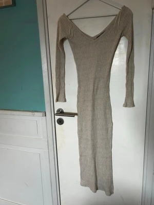 Beige ribbad långklänning - Långklänning i beige ribbat material med v-ringad hals och långa ärmar. Klänningen har en figurnära passform och är tillverkad i ett stretchigt tyg som känns mjukt mot huden. Perfekt för dig som gillar en enkel men snygg stil.
