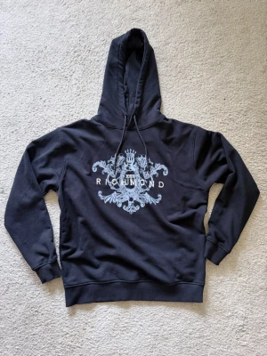 Svart hoodie med tryck från Richmond - Snygg svart hoodie från Richmond med stort vitt tryck på bröstet som har ett detaljerat emblem och texten 'Richmond'. Klassisk modell med huva och dragsnören, ribbade muddar vid ärmar och nederkant. Perfekt för en avslappnad och trendig stil.