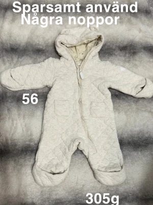 Beige overall med öron från Newbie - Mysig beige quiltad overall från Newbie med huva och söta öron. Overallen har dragkedja framtill, två fickor och mjukt innerfoder med djurmönster. Perfekt för kyliga dagar och enkel att ta på. Passar små barn.🌸 Sparsamt använd. Några noppor. Storlek 56 