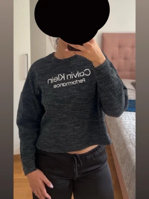 Mörkgrå Calvin Klein cropped sweatshirt - Säljer en mörkgrå cropped sweatshirt från Calvin Klein Performance med vit logga framtill. Tröjan har rund halsringning, långa ärmar och en avslappnad passform. Perfekt för en sportig och chill look.
