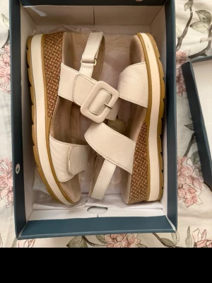 Vita platåsandaler med stor spänne från Scorett - Snygga vita sandaler från DASIA, Scorett med platåsula och stor dekorativ spänne. Ovandelen är i slätt material och sulan har ett flätat mönster på sidan. Justerbar rem runt hälen och breda justerbara remmar över foten. Inte använda många gånger men tyvärr lite mörka på innersulan.