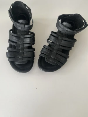 Svarta gladiatorsandaler i läderimitation - Säljer ett par svarta gladiatorsandaler med flera remmar och öppen tå. Sandalerna har en robust sula och justerbart spänne vid ankeln. Tillverkade i läderimitation och passar perfekt till sommarens outfits.