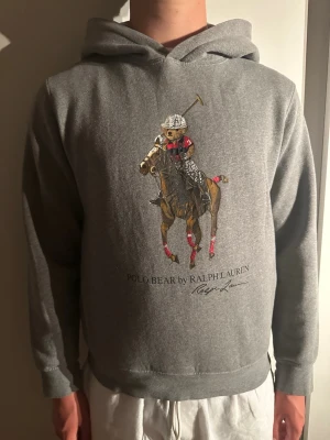Grå hoodie Polo Bear Ralph Lauren - Snygg grå hoodie från Ralph Lauren med Polo Bear-tryck framtill där björnen rider häst i poloutstyrsel. Klassisk huva och ribbade muddar. Perfekt för dig som gillar streetwear och vill ha något bekvämt men ändå stilrent.