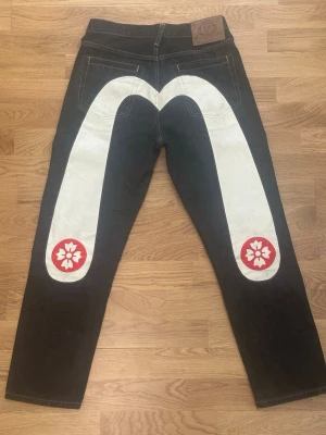 Svarta Evisu jeans med vit grafik - Evisu svarta jeans med stor vit grafisk båge och röda blommor på bakstycket. Jeansen har raka ben och klassisk femficksdesign. Materialet är kraftig denim från Japan och de har en läderpatch med Japansk illustration i midjan.