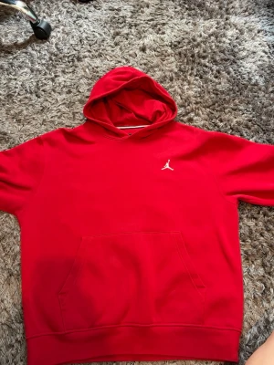 Röd Jordan hoodie - Snygg röd hoodie från Jordan med klassisk vit Jumpman-logga broderad på bröstet. Hoodien har stor känguruficka framtill och en rymlig huva. Perfekt för dig som gillar streetwear och sportig stil.