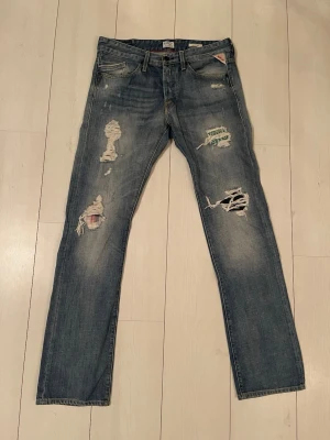 Blå slitna Replay jeans med hål - Säljer ett par blå jeans med tydliga slitningar och stora hål på benen för en riktigt edgy look. Jeansen har klassisk femficksmodell, raka ben och detaljerade slitningar framtill. Perfekta för dig som gillar streetstyle och vill sticka ut. Storlek W33 L34