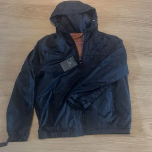 Svart vindjacka med rutigt foder - Snygg svart vindjacka med huva och dragkedja framtill. Insidan har ett klassiskt brunt, vitt och vinrött rutigt foder. Jackan har två fickor med dragkedja och justerbara muddar vid ärmsluten. Perfekt för blåsiga dagar och enkel att matcha.