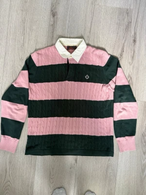 Morris Stockholm Kabelstickad Rugbytröja / Polo – Storlek L - Snygg och klassisk kabelstickad rugbytröja från Morris Stockholm i rosa och mörkgröna ränder med vit krage och broderad logga på bröstet. Riktigt skön modell med knäppning i halsen som ger en klockren preppy-/collegestil. 