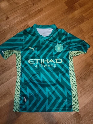 Manchester City målvaktströja grön - Snygg Manchester City målvaktströja i grönt med grafiskt mönster och gula detaljer på sidorna. Puma-logga på bröstet och Etihad Airways som sponsor. På ryggen står det 'EDERSON M' och nummer 31. Perfekt för fotbollsträning eller match.