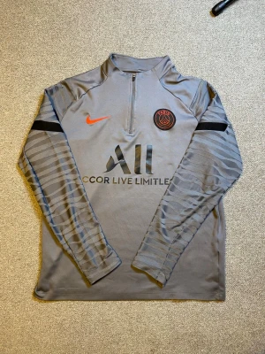 Grå PSG träningströja från Nike - Snygg grå långärmad träningströja från PSG med halv dragkedja, svarta och orange detaljer samt klubbmärke på bröstet. Tillverkad i funktionsmaterial som passar perfekt till fotbollsträning eller gym. Diskreta mönster på ärmarna och svart logga på bröstet.