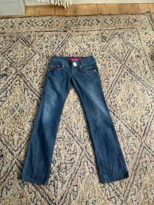Blå bootcut jeans med röda detaljer - Snygga blå bootcut jeans med klassisk femficksmodell och röda detaljer på fickorna. Jeansen har normal midja och kontrastsömmar. Tillverkade i ett mjukt denimtyg som ger en bekväm passform. Perfekta för dig som gillar en tidlös jeansstil.