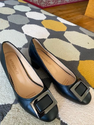 Svarta pumps med stor dekorativ spänne - Stilrena svarta pumps i skinnimitation med chunky klack och stor fyrkantig metallspänne framtill. Skon har rundad tå och beige innersula med diskret mönsterkant. Perfekta för dig som gillar klassisk men trendig look.