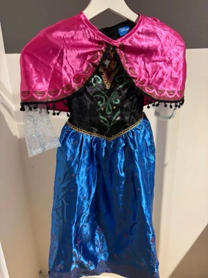 Disney Anna maskeradklänning Frost - Maskeradklänning inspirerad av Anna från Frost. Klänningen har svart överdel med gröna och rosa detaljer, blå glansig kjol med snöflingor, transparenta glittriga ärmar och en avtagbar rosa cape med dekorativa bollar. Perfekt för cosplay eller maskerad!